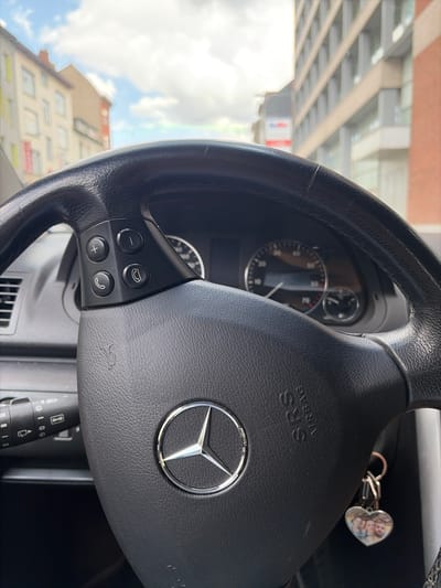 The image is taken from inside a car, looking at the steering wheel, dashboard, and through the windshield. The steering wheel is black leather and has several buttons on the left side, including volume controls and call buttons. The Mercedes-Benz logo is visible on the steering wheel. To the right of the steering wheel, a portion of the dashboard is visible, including a speedometer that reads approximately 60. A keychain with a heart-shaped charm containing three small photos hangs from the steering column.
Outside the car, buildings are visible. On the left, there are multi-story buildings with windows, one of which has yellow accents. On the right, a more modern building with a grid-like facade is visible. The sky is partly cloudy.
The scene is located on a street in the city of Schaerbeek - Schaarbeek, Belgium. The presence of multi-story buildings and the architecture suggest an urban environment. The cars and buildings are consistent with a European city. The weather appears to be partly sunny.