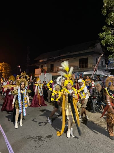 This nighttime image depicts a street procession occurring in Kedungbanteng, Indonesia, specifically in an area identified by a building sign as "Desa Kali, RT 12 RW." The scene is illuminated by artificial light sources against a dark sky.

In the foreground and midground, a group of at least five individuals are actively participating, dressed in elaborate, culturally-inspired costumes. The central figure wears a predominantly gold and black outfit, featuring a large headpiece adorned with white and yellow feathers and yellow floral garland-like structures, complemented by white boots. To their immediate left, another participant is visible in a gold and black costume with a prominent gold headpiece and white boots; this costume incorporates what appear to be numerous packaged goods (e.g., snack wrappers) as decorative elements. Further left, two individuals are clad in flowing maroon gowns with gold shoulder accents and large gold headpieces. A partial view of a fifth participant in a brown, animal-like costume is visible on the far right.

Along the right side of the street and in the background, a crowd of spectators, comprising adults and children, observes the procession. A dark-colored scooter with the visible license plate "2364 NJ" is parked among the spectators on the right. In the background, a two-story building with a dark, sloped roof and a balcony with black railings is prominent. Multiple red and white Indonesian flags are displayed on and around this building. Trees with visible foliage are situated on the far left and far right edges of the frame. The procession takes place on an asphalt road.