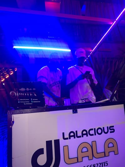 The image shows two individuals, likely DJs, at a nighttime event in Shendam, Nigeria. Both are wearing white t-shirts and hats, suggesting a casual, party atmosphere. The DJ on the left is interacting with a laptop and equipment, while the DJ on the right is looking at a mobile phone. The area is illuminated by vibrant blue and pink neon lights, typical of a club or outdoor entertainment venue. A prominent banner on the front of the DJ booth displays the text "LALACIOUS LALA" and a phone number, indicating branding for the event or performers. The overall impression is one of a lively, modern social gathering.