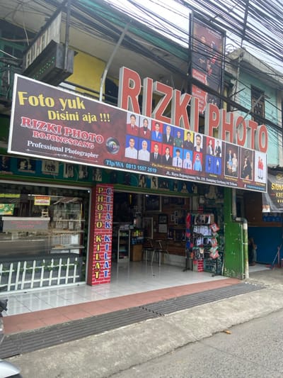 The image shows the storefront of a photography studio named Rizki Photo, located at JL. Raya Bojongsoang No. 223, Sampling, Gerbang Sit. Tekan, Bojongsoang, Indonesia, according to the sign. The red, yellow, and white sign is partially obscured by wires and reads "Foto yuk Disini aja!! Rizki Photo Bojongsoang Professional Photographer Studio Tlp/WA 0813 1557 2619." There are many framed photographs of people, some in formal attire, displayed in the storefront windows. Several chairs are visible inside the studio. The sidewalk in front of the studio is paved with concrete and has a ramp leading to the street. A motorcycle is parked on the sidewalk in front of the store. The storefront is decorated with green painted brick and green plants. The building's exterior is grey. The studio is located on a busy street, with many other buildings and businesses nearby. Wires and electrical lines are strung across the street. The image was likely taken on a sunny day.