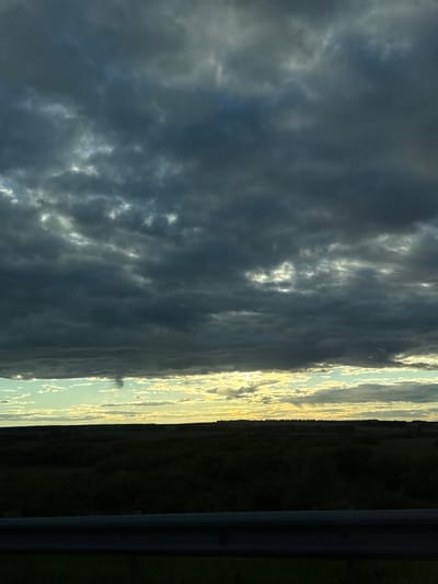 A dark, stormy sky looms over a rural landscape. In the distance, a thin funnel cloud extends from the clouds towards the horizon, indicating a potential tornado. The foreground is dominated by dark, shadowy trees and vegetation, with a metal guardrail running horizontally across the bottom of the image. The sky is a mix of dark gray clouds with patches of bright light breaking through, creating a dramatic contrast. The location is identified as Kuralovo, Russia.