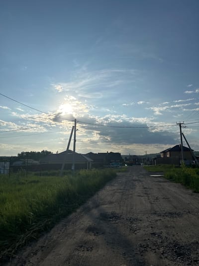 The image depicts a rural dirt road leading into a residential area under a partly cloudy sky. The sun is partially obscured by clouds, casting rays of light. Power lines stretch across the scene, supported by utility poles. On either side of the road, there is tall green grass and some vegetation. Several houses with visible roofs are situated along the road. In the background, a treeline is visible, and a slight incline suggests a gentle hill. There are no people or animals visible in the frame. The overall impression is that of a quiet, sunlit day in a village setting. The location is the village of Estachi, Russia.