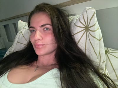 A medium shot captures a woman lying in bed, her head resting on a pillow with a geometric pattern. She has long, dark hair that frames her face and falls over her shoulders. Her eyes are open and looking slightly to the left, and her lips are slightly parted. She is wearing a light green top and a silver cross necklace. The background shows a wall and what appears to be a headboard. The lighting is dim, suggesting an indoor setting at night. The overall scene appears to be a private, relaxed moment. The location of Мосметаллоконструкция Онт, Russia is not discernible from the image.