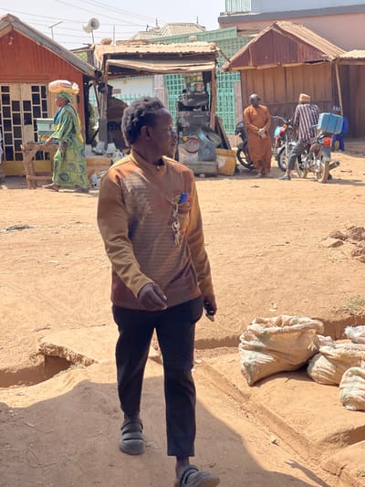 The scene depicts an unpaved commercial area in Chikaji, Nigeria, under daylight conditions. In the foreground, a dark-skinned male, wearing a brown patterned long-sleeved shirt, dark pants, and dark sandals, walks rightward. A pen and keys are clipped to his left chest pocket. To his right, several beige sacks are piled on an elevated dirt section. In the midground, a woman in a green and yellow dress with a white headwrap carries a white sack on her head, moving left past a reddish-brown wooden stall. Further right, two men are present near motorcycles: one in an orange garment stands next to a red motorcycle, while another in a striped shirt and hat is near a motorcycle fitted with a blue delivery box. The background features various simple structures with corrugated metal roofs, including one with loudspeakers. The ground consists of dry, reddish-brown dirt.