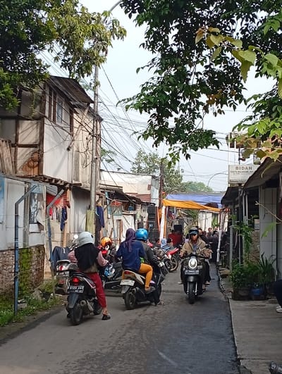 A narrow street scene is bustling with motorcycle traffic. Several riders are positioned mid-street, with their vehicles oriented towards the camera. The buildings on either side are modest and closely spaced, with some featuring visible laundry lines and basic construction. An overhead tarp structure and gathering of people hint at a possible event in the distance. Overhead, tangled power lines weave amidst trees, adding to the dense urban landscape.