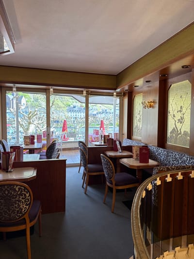 The image shows the interior of a restaurant in Cochem, Germany, with tables and chairs positioned near large windows offering a view of the city. The restaurant's interior features decorative panels on the ceiling with floral designs and ornate lamps. Tables are set with menus, and some are covered with patterned cloths. Outside, the windows reveal Cochem's buildings, trees, and the Moselle River. A red umbrella is visible at an outdoor seating area. The scene appears calm and unoccupied at the moment of the photo.