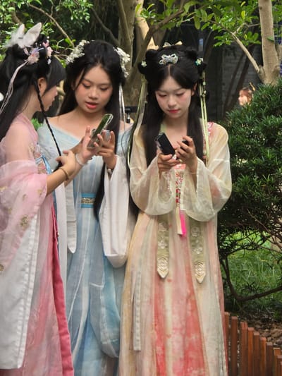 Three young East Asian women, dressed in traditional Hanfu-style attire, stand outdoors. The woman on the left wears a pink robe with gold embroidery. The woman in the middle wears a light blue robe with white trim and a blue sash. The woman on the right wears a pale yellow and pink robe with white lace trim and floral embroidery. All three women are looking down at their mobile phones, with their fingers interacting with the screens. They are standing in a garden or park-like setting with lush green trees and foliage in the background, and a wooden fence in the foreground. The light suggests it is daytime. The presence of traditional clothing and the style of the hairstyles suggest a cultural event or photo shoot, possibly in a scenic location known for its natural beauty, such as Xihu District in China.