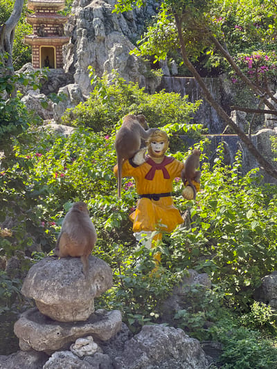 In Ninh Ích, Vietnam, a collection of monkeys interacts with a statue of Sun Wukong, the Monkey King, in a lush, rocky environment with a pagoda in the background. Three monkeys are visible in the foreground, one perched on a rock formation, another on the statue's shoulder, and a third clinging to the statue's arm. The statue is adorned in a yellow robe with a red sash and a black belt. In the midground, the statue stands among dense green foliage and pink flowers, with a cascading waterfall visible behind it. In the background, a multi-tiered pagoda rests against a rocky cliff face, with two more monkeys sitting on its roof. The overall scene depicts a harmonious coexistence between wildlife and cultural elements in a vibrant natural setting.