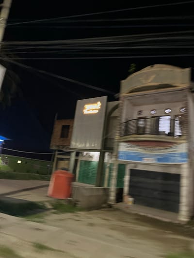 The image depicts a blurry nighttime street scene in Bundar, Indonesia. In the foreground, a concrete sidewalk is visible, with a dark, wet area suggesting recent rain or a ditch. To the left, a green building with illuminated windows can be seen. Further back, an orange water tank stands next to a building under construction.
The main focus of the image is a multi-story building on the right. The lower level appears to be a shop with a large, dark shuttered entrance. Above this, a section with a blue and white banner is visible. The second floor has a balcony with a dark railing and several windows. The top of the building features an arched architectural element.
Above the buildings, a dense network of power lines stretches across the dark sky. A brightly lit sign with orange lettering is affixed to a facade on the left side of the street, partially obscuring the building underneath. The overall impression is of a modest urban environment at night, characterized by a mix of commercial and residential structures.