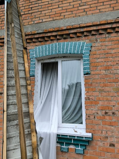 The image depicts a partial view of an exterior brick wall of a building in Pysarivka, Ukraine. The wall is constructed from reddish-brown bricks with grey mortar lines. A white polyvinyl chloride (PVC) framed window with two vertical panes is centrally positioned. The left pane of the window is ajar, with a sheer white curtain partially extending outwards, indicative of airflow. A denser, light grey or blue curtain is visible behind the closed right pane. The window is adorned with decorative brickwork painted in a teal or blue-green color, featuring vertical white stripe patterns above and below the window. Above the window, a horizontal concrete lintel and a decorative course of projecting bricks are visible within the red brickwork. To the immediate left of the window, a temporary construction structure, resembling a makeshift ladder or scaffolding, leans against the wall. This structure consists of two parallel wooden planks with horizontal metallic or composite steps. The wall section directly behind this apparatus is covered with a corrugated grey sheet material. The scene is illuminated by diffuse natural light. No individuals are present.