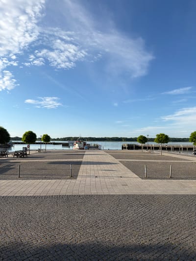 The image depicts a waterfront promenade in Ribnitz-Damgarten, Germany. The scene is oriented vertically, with the sky occupying the left portion and the promenade and water on the right. The sky is a vibrant blue, with scattered white clouds, suggesting fair weather and daytime. The promenade is paved with two different materials: a section of light-colored square tiles and a section of dark cobblestones. Several trees, pruned into rounded shapes, are planted along the promenade, with benches placed between them.
In the background, a body of water is visible, with a tree-lined shore on the opposite side. A white boat with a red stripe and the text "Schiffsausflugshafen" (meaning "Excursion Boat Harbor" in German) is docked near the promenade. The presence of the boat and the harbor text clearly indicates a location related to water transportation and tourism. The time of day appears to be late morning or early afternoon, given the angle of the shadows cast by the trees and posts on the promenade. The overall atmosphere is peaceful and suggests a pleasant day for a stroll or relaxation by the water. There are no people visible in the image.