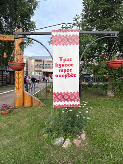 The image shows a banner with red and white ethnic patterns and text in Russian hanging from a metal archway supported by wooden posts. The banner reads "Трос купос-трос иорвёс". In the background, there is a building decorated with colorful flags. A group of people are gathered in front of the building. There are potted plants with flowers hanging from the archway and on the ground. The scene is set outdoors with a grassy area and trees. The provided coordinates place the image in the village of Kukuy, Russia.