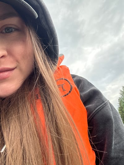 A close-up, chest-up portrait captures a young woman outdoors. She wears a dark hooded sweatshirt and a bright orange life vest, partially obscured by her long, light brown hair. The life vest has a circular emblem with a black border and a black letter "T" inside. Her face is turned towards the camera, with her left eye, nose, and mouth visible. The background consists of a cloudy, grey sky and a hint of green trees on the right side of the frame. The setting appears to be a natural environment, possibly near water, given the life vest. There is no identifiable information suggesting the location is Shul'dikha, Russia.