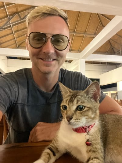 An adult male and a tabby cat are depicted in a close-up image taken indoors or within a semi-open structure in Uluwatu, Indonesia. The male, positioned on the left, has short blond hair, is wearing aviator-style sunglasses with dark frames and light-tinted lenses, and a grey t-shirt with horizontal darker stripes. He is smiling directly at the camera. A light brown and white tabby cat, wearing a red collar with a small red bell, sits on a wooden table in front of the male, looking slightly off-camera to the right. The background reveals white structural beams supporting a thatched or woven natural material ceiling. Out-of-focus wooden furniture is visible in the distant background.