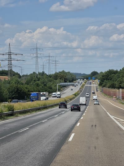 The image depicts a sunny day on a multi-lane highway with traffic. In the foreground, the road surface is visible, with lane markings. Several cars and vans are driving on the highway. In the middle ground, more vehicles are visible, including trucks. In the background, a dense line of trees borders the highway, and tall electrical pylons are prominent against a blue sky with white clouds. The scene is likely on a highway near Ebernhahn, Germany, given the presence of German license plates on some vehicles and the general landscape, which is consistent with the region. There are no people clearly visible in the image. The overall impression is one of typical highway traffic flow in a rural or semi-rural setting.