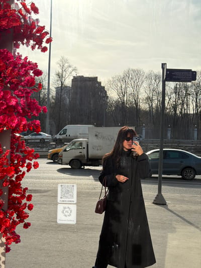 The image depicts an outdoor urban scene in Perepechino, Russia, under bright daylight with visible shadows. A female individual, dressed in a long, dark coat and dark sunglasses, is positioned centrally, holding a smartphone in her right hand as if taking a selfie. She has long, dark hair and carries a small, dark red handbag over her left shoulder. The individual appears to be reflected in a large, vertical reflective surface, possibly a window, which shows streaks and glare. On this reflective surface, two white square decals are visible: one with a QR code and the text "FLOWERS.GERBERA," and another below it featuring a stylized floral logo and "Flowers Gerbera" text.
To the immediate left, a dense cluster of vibrant red artificial flowers extends into the frame. Beyond the reflective surface and the individual, a multi-lane asphalt road is visible with several vehicles, including a white cargo van, a light-colored pickup truck, a yellow sedan (possibly a taxi), a dark grey sedan, and a white sedan in the distance. Further into the background, a street sign on a dark pole points right, displaying Cyrillic text indicating "MVD Perepechino" (Ministry of Internal Affairs Perepechino) and "GK Peredelkino." Bare deciduous trees line the far side of the road, and multi-story buildings, including one large, older-style structure, are visible against a bright, hazy sky.