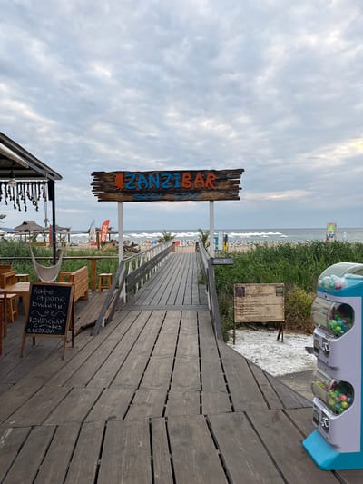 The image depicts a wooden walkway leading to a beachside establishment named "ZANZIBAR BEACH STYLE" in Lozenets, Bulgaria. The sky is overcast with clouds. To the left of the walkway, there is an outdoor seating area with wooden tables and chairs, a hammock, and a chalkboard menu. The menu is written in Bulgarian and appears to list coffee, paninis, and non-alcoholic cocktails. To the right of the walkway, a gumball machine is visible. Beyond the walkway and seating area, the beach is lined with rows of white umbrellas and lounge chairs. The sea is calm. Vegetation, possibly reeds or tall grass, grows between the walkway and the beach. In the background, several flags are visible, including one with the "APEROL" logo. The overall scene suggests a relaxed, casual beach bar or restaurant setting.