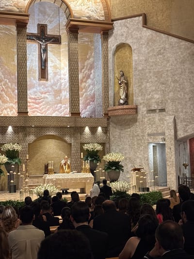 An aerial shot captures an interior scene within a church, likely in San Pedro Garza García, Mexico, during a religious ceremony. In the foreground, a congregation is seated, their backs to the camera, filling the pews. They are dressed in formal attire, with women in dresses and men in suits, suggesting a special occasion such as a wedding or a baptism. The middle ground features an altar area, where a priest in gold vestments is officiating, holding a chalice. The altar is adorned with floral arrangements and candles. The background reveals the ornate architecture of the church, including a large crucifix hanging from the ceiling, columns, and decorative murals depicting clouds. The lighting is dim, with candles providing a soft glow, contributing to a solemn atmosphere.