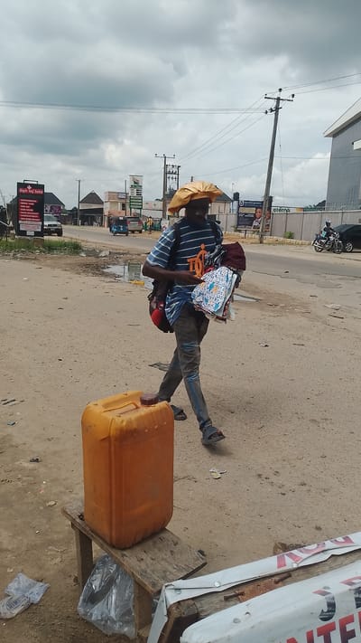 A man walks along a dirt road in Warri, Nigeria, carrying a bundle of clothing and a red object under his arm. He wears a blue and white striped t-shirt, dark pants, and dark sandals. A yellow jerrycan sits on a wooden stool to his left. In the background, buildings, power lines, and a few vehicles, including a white van and a blue auto-rickshaw, are visible. The sky is overcast.