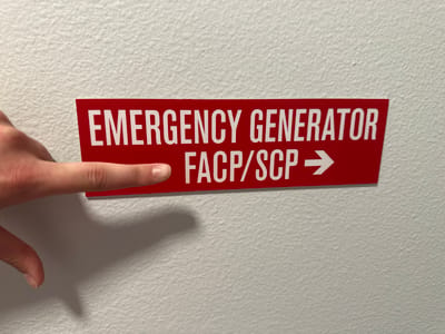 A person's index finger points to a red sign with white lettering that reads "EMERGENCY GENERATOR FACP/SCP ->" on a textured white wall. The sign is rectangular with a slightly angled right edge. The finger is positioned over the word "FACP". The background is a plain, textured white wall. The image appears to be taken indoors, possibly in a building in Lynnwood, United States, given the context provided, though the location is not explicitly visible in the image itself.