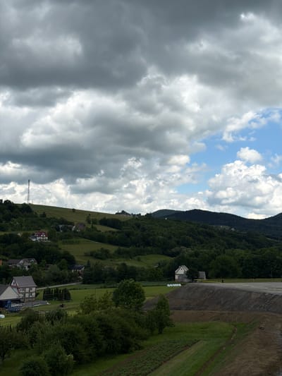 This is a landscape photograph taken on a cloudy day in the city of Janowice, Poland. The image features rolling green hills covered in dense trees and dotted with scattered houses. In the foreground, a construction site is visible, with a large pile of dirt and a paved area. The sky is filled with dramatic, dark clouds interspersed with patches of blue sky and brighter, lighter clouds. A tall telecommunications tower stands on a distant hilltop. There are no people visible in the frame. The scene depicts a rural or semi-rural environment undergoing some form of development or renovation.
