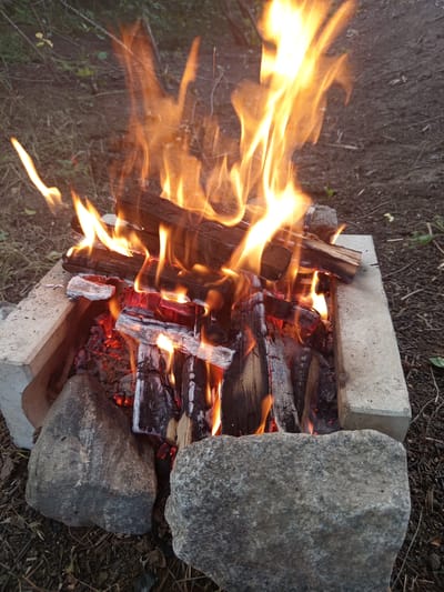 A campfire is burning in a makeshift fire pit constructed from concrete blocks and stones. The fire consists of several pieces of wood that are actively burning, with flames reaching upwards. The surrounding ground is covered in dirt, dry grass, and twigs. The image appears to be taken outdoors, likely in a natural or semi-natural setting. There are no people visible in the image. The location in Vilshany, Ukraine, is not discernible from the visual information provided.