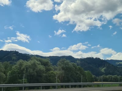 The image depicts a scenic view of rolling green hills and mountains under a bright blue sky with fluffy white clouds. The foreground shows a metal guardrail and a glimpse of a road, suggesting the photo was taken from a vehicle. Lush green trees cover the lower slopes of the mountains, transitioning to darker, denser pine forests higher up. Patches of lighter green indicate clearings or meadows on the mountainside. The overall impression is of a natural landscape, likely in a rural area. The location is identified as Sankt Marein-Feistritz, Austria.