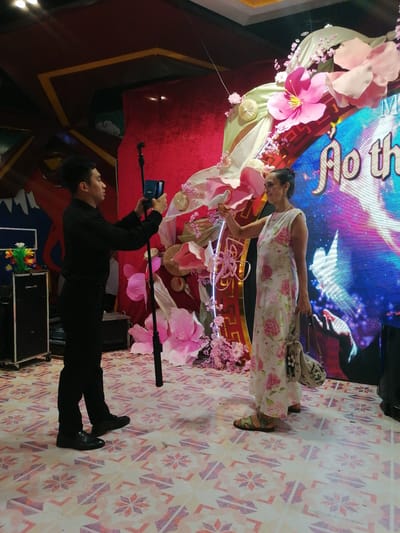 The image captures a scene in Hong Gai Ward, Vietnam, likely at a promotional event or party. A man, dressed in a black shirt and pants, is taking a photo with a smartphone on a tripod. He is facing a woman in a floral dress who is holding a white dove. The background features a large decorative display with pink flowers, a screen with text on it, and red fabric. The floor has a geometric pattern. A black equipment case is on the side of the frame.