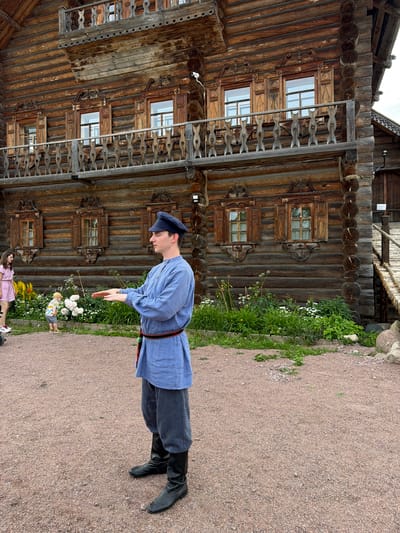 A man in traditional clothing stands in front of a wooden building in Dnp Ryzhiki, Russia. He is wearing a blue tunic, striped pants, and black boots, and a blue cap. His hands are held out in front of him, palms up, as if gesturing. In the background, a woman in a pink dress and a small child in a yellow hat are visible. The building appears to be an old wooden house with intricately carved window frames and a balcony. The ground is covered in gravel.