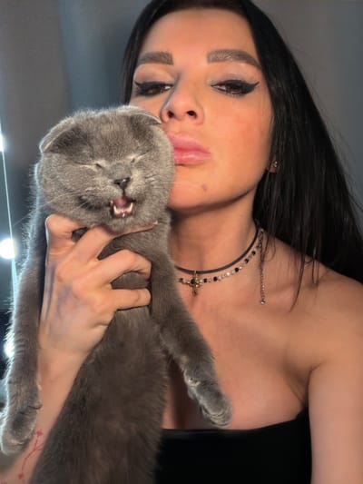 A medium-shot portrait depicts a female individual holding a solid grey Scottish Fold cat indoors, with the scene identified as Svkh Maslovskiy 1 Otdeleniye, Russia. The woman, positioned on the right, possesses long, dark hair and prominent make-up, including winged black eyeliner and filled eyebrows. Her lips are puckered into a 'kissing' gesture, directed towards the cat's head. She wears a black strapless garment and two layered necklaces: an upper black choker with a small golden cross pendant and alternating clear and black beads, and a lower, thinner metallic chain. A small stud earring is visible in her right ear, and a faint red, possibly floral, tattoo is present on her left forearm. Her right hand secures the cat in a vertical orientation. The cat, centrally positioned, exhibits squinted eyes and an open mouth, revealing its tongue and upper and lower teeth in what appears to be a pronounced vocalization or expression. The background is uniformly light grey and out of focus, with several blurred bright light sources visible on the left side.