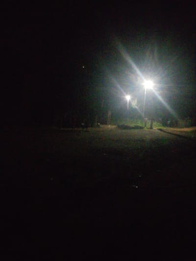 A nighttime scene depicts an outdoor area with two streetlights illuminating a dimly lit path or road. The streetlights are bright and cast beams of light that create lens flares and illuminate the surrounding environment. In the background, trees and vegetation are visible, suggesting a park or a natural setting within or near Kagoma, Nigeria. The ground appears to be covered in gravel or dirt. There are no visible people or animals in the image. The overall impression is one of quiet and solitude, with the artificial light from the streetlights being the primary source of illumination in the darkness.