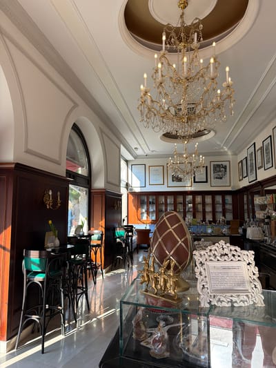 Here is a detailed description of the image:
The image captures the interior of what appears to be a high-end café or confectionery, possibly located in the city of Grad Opatija, Croatia, based on the writing on a display case inside. The perspective is taken at an angle, looking upwards towards the ceiling, which features a large, ornate chandelier, and diagonally towards the back of the room.
On the right side of the image, there's a display case filled with sweets and cakes. A white, decorative frame is visible. In front of it is a brown, sphere-shaped object and a gold-colored statuette of running rabbits.
Behind the display case, there are shelves lined with glass jars and bottles, implying a selection of ingredients or goods for sale.
The ceiling is adorned with a golden-colored oval design and detailed moldings.
On the left side, there's an arched window or doorway, through which a glimpse of the outside world can be seen.
In the middle of the image, there are several black chairs stacked or lined up, possibly for customer seating. There are also dark wooden cabinets or counters with vintage-style sconces.
The setting appears to be a blend of elegance and old-world charm.
