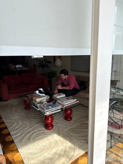 A mid-day interior view captures a living space in Porto Alegre, Brazil, observed through an opening partially obscured by a white roller blind and a white vertical frame. A male individual, wearing a reddish-pink long-sleeved shirt and dark trousers, sits on a light brown fabric sofa positioned on the right side of the frame. He holds a small, dark object in his right hand and appears engaged with a black laptop on a central glass-top coffee table. This table features four glossy red, turned-wood legs and rests on a cream-colored rug with subtle wavy patterns. The table surface is covered with numerous stacked books and magazines, including one identifiable as "Atlas Ambiental de Porto Alegre," alongside a small white box bearing an 'H' logo and various cables. To the left, a large red fabric sofa is visible. The flooring visible beyond the rug is a geometric parquet wood pattern. In the background, a dark wooden dining table, potted plants, and wall-mounted art, including a large dark rectangular piece and a white spherical decorative object, are discernible. Natural light from the left illuminates a section of the rug.