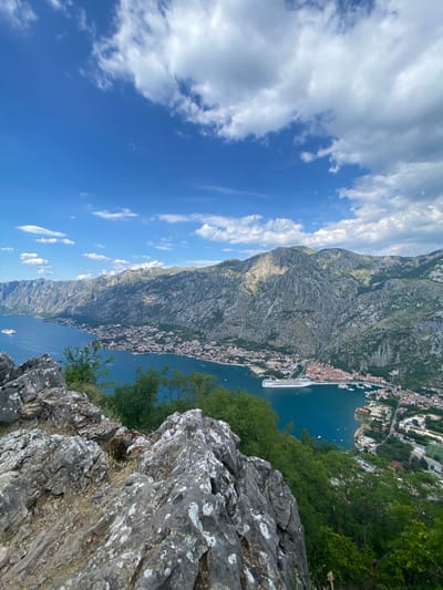 The image depicts a panoramic view of the Bay of Kotor, likely from a viewpoint above the town of Škaljari, Montenegro. In the foreground, rugged, weathered gray rocks dominate the lower left of the frame, interspersed with dry grasses and green foliage. Beyond the rocks, a dense canopy of green trees extends down a slope towards the water.
The bay itself is a deep, tranquil blue, reflecting the bright sky above. A large white cruise ship is moored in the bay, near a town that stretches along the coastline. The town features red-tiled roofs and appears to be nestled at the base of towering, steep, rocky mountains. These mountains, etched with shadows and highlights from the sun, form a dramatic backdrop, extending as far as the eye can see. The sky is a vibrant blue, dotted with scattered white, fluffy clouds. The overall impression is one of a majestic natural landscape juxtaposed with a human settlement. There are no people visible in the image.
