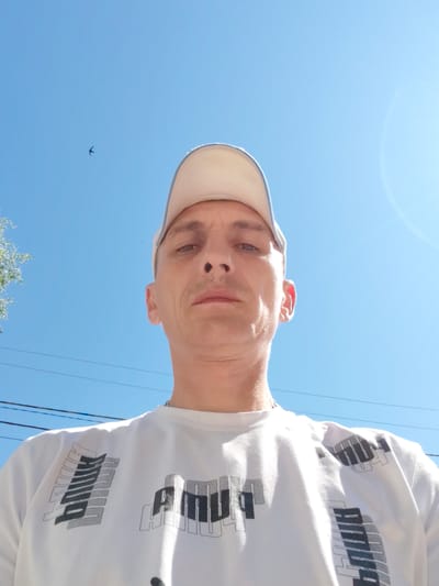 A low-angle, medium shot captures a light-skinned male, appearing to be of Caucasian descent, wearing a white t-shirt with a graphic print and a light-colored baseball cap. He is outdoors, with a clear blue sky dominating the background, suggesting a daytime setting. A small bird is visible in flight against the sky, and tree branches are present on the left side of the frame. Electrical wires are stretched across the background. The man's face is the central focus, with a neutral expression. His gaze is directed upwards, towards the camera. The overall lighting suggests bright sunlight. The image's perspective and backdrop indicate an outdoor urban environment, potentially in the city of Bor, Russia.