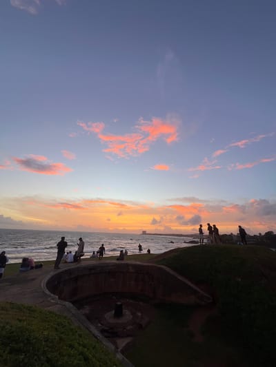 This media file depicts a sunset scene at Galle, Sri Lanka. Several people are visible, silhouetted against the colorful sky and sea. They are standing and sitting along a curved, paved walkway that borders a grassy embankment. The sea is calm, with gentle waves lapping at the shore. The sky is filled with dramatic clouds tinged with orange and pink, indicating the time of day is sunset. The overall atmosphere appears to be peaceful and contemplative, with people enjoying the scenic view. The location cues, such as the architecture and the presence of a historical structure with a circular opening, suggest a place of historical significance or a tourist attraction. There is no visible text in the image.