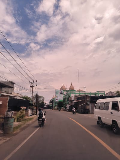 The image captures a street scene in Keudejunieb, Indonesia, under a cloudy sky with a distinct reddish-pink hue, suggesting either dawn or dusk. The asphalt road features a solid yellow line down its center.
On the left side of the road, two blue-green barrels are visible, one clearly labeled "SOLAR" (likely for diesel fuel). Behind them are various residential or commercial buildings, some with corrugated roofs, and a tall utility pole with a dense network of overhead wires stretching across the frame. A person wearing a white shirt and black backpack is riding a motorcycle, with a passenger behind, heading away from the viewer.
Further down the street, on the right, stands a prominent building characterized by its green and white facade and two golden domes, resembling a mosque or Islamic education center. A large banner hangs from this building, displaying text that reads "PENERIMAAN SANTRI BARU" (New Student Enrollment) and includes phone numbers: "0822 1092 5190" and "0822 3247 2541". Several other buildings and some vegetation line the sides of the road. A white minivan is partially visible on the right, moving in the same direction as the motorcycle in the foreground. Another motorcycle is seen further down the road, and what appears to be a black pickup truck is parked on the side.
The scene depicts a typical daily activity of commuting and local life in a town or city.