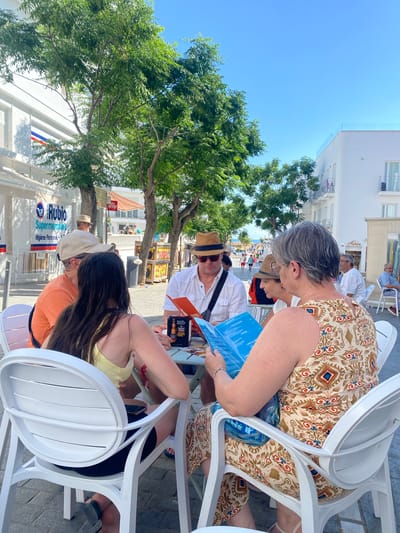 A group of four people are seated at an outdoor cafe table on a sunny day in Conil de la Frontera, Spain. The cafe appears to be located on a pedestrian street lined with white buildings and trees.
In the foreground, two women are seated. The woman on the right, with graying hair pulled back, wears a patterned sleeveless dress and is looking down at a blue menu. The woman on the left, with long dark hair, is wearing a yellow tank top and is looking away from the camera.
Behind them, two men are seated. The man on the left, wearing an orange t-shirt and a beige baseball cap, faces the women. The man on the right, wearing a white button-down shirt and a straw hat, is looking at a red menu and appears to be speaking or interacting with the people across from him. A bottle of Estrella beer and a small box are on the table.
In the background, other cafe patrons and pedestrians are visible. Buildings with white facades and balconies are present on both sides of the street. A sign for "Rubio Supermercados" is visible on the left. The overall impression is a relaxed, daytime scene at a cafe.