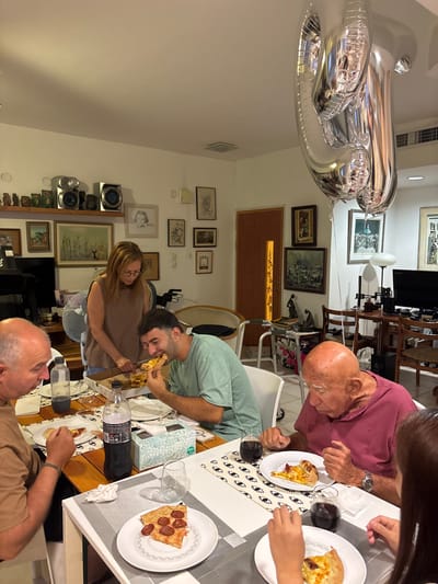 This image depicts an indoor family gathering in a residence in Bnei Dror, Israel, centered around a meal of pizza. Five individuals are present around a light-colored wooden dining table. In the left foreground, a balding man in a light brown shirt is seated, observing the table. To his right, a younger man in a light green t-shirt is actively consuming a large slice of pizza. Behind these two, a woman with long brown hair, wearing a striped sleeveless top, stands and uses a knife to cut pizza within a cardboard box on the table. In the right foreground, an elderly bald man in a maroon polo shirt is seated, looking down at a slice of pizza on his white plate, with a glass of dark liquid beside it. Partially visible to his right is another person's arm, also with a plate of pizza.

The table is furnished with white plates, a clear water bottle, a dark soda bottle with Hebrew labeling, a light blue tissue box, and patterned placemats. Two large, silver, reflective foil balloons, shaped like numbers, float near the ceiling in the upper right, indicative of a celebration. The background features white walls extensively decorated with framed artwork of various sizes and styles. A wooden shelf on the left wall holds decorative owl figurines and a silver stereo system. A dark television or monitor is visible below the shelf. In the background, a light brown wooden door with a vertical glass panel is visible, along with a silver-framed wheeled walker. To the far right, a desk with a computer monitor and wooden chairs is present. The room is illuminated by warm, artificial lighting.