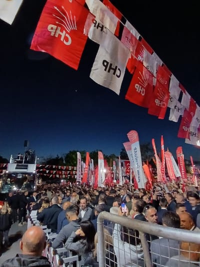 A large crowd of people are gathered at a political rally in Silivri, Türkiye, at the coordinates 41.071, 28.244. The crowd is standing in front of a fence, with red and white flags with the letters "CHP" visible above the crowd. The flags are hanging from a large truck that is parked in the background. There are many people in the crowd, some of whom are holding up their phones to record the event. The event is taking place at night, and the street is lit by streetlights. The rally appears to be a public gathering in support of the CHP party.
