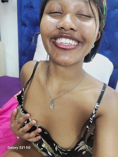 A woman with dark hair and a headband is smiling widely with her eyes closed. She has a nose ring and is wearing a silver necklace with a leaf-shaped pendant. The woman is wearing a black top with a floral pattern, and her hand is resting on her chest. The background includes a blue textured surface and a glimpse of pink fabric. "Galaxy S21 5G" is visible in the bottom right corner of the image.