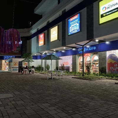 The scene depicts an illuminated outdoor commercial plaza at night in Tumbaco, Ecuador. A multi-story building occupies the background, featuring a light-colored upper section and a darker lower section accented with a blue band.
On the upper facade, several illuminated signs are visible: from left to right, "MUNDO MELLAS," a large digital display advertising "OMNIA ESENCIAL" with "Triple Esencial para tu Bienestar! 2x $25," and "RED ACTIVA Western Union." Below these, ground-level storefronts with large glass windows are visible, including one branded "OMNIA ESENCIAL" displaying merchandise like shoes. To the far left, a "PYCCA" sign is also visible above another storefront.
The plaza ground is paved with rectangular gray interlocking pavers. In the mid-ground, several sets of white outdoor tables and chairs are arranged. Two of these sets are positioned under large green and white striped market umbrellas. To the left, a small, white kiosk or stand is present.
Four individuals are seated at a table under the left-most umbrella, facing each other, engaged in an unseen activity or conversation. No specific actions are discernible beyond their seated posture. Green foliage, comprising small trees and shrubs, is planted in raised beds along the base of the building's storefronts and near the seating areas. To the far left, an overhead decorative structure made of purple and red streamers hangs from the building. The entire area is well-lit by artificial lights integrated into the building's overhangs and other fixtures.
