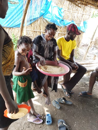 The image depicts a scene in Otulu, Nigeria, under a makeshift shelter with a blue tarp and thatched roof. The ground is dirt, with scattered debris and several pairs of sandals. Sunlight streams in, suggesting it's daytime, possibly hot and humid.
The central figure is a woman wearing a black and gray patterned top, smiling broadly as she holds a pink plastic basin filled with what appears to be rice or grain. To her left, a young child, a boy, is held by his mother, wearing a green skirt with an orange pocket. The child looks engaged with the basin, his expression a mix of curiosity and slight apprehension. He wears a cross necklace.
To the right of the woman is a man wearing a bright yellow t-shirt and a red and black plaid baseball cap. He is seated on a wooden bench, his arms resting on his legs. His gaze is directed downwards, away from the camera. His dark pants are visible.
Another woman is partially visible on the far left, wearing a yellow and black patterned garment. She is holding an orange and white plate. Her face is not visible.
The atmosphere seems informal and communal, with people gathered together, possibly sharing food or engaged in a task related to the grain. The woman's joyous expression suggests a positive social interaction, while the child's stance indicates participation in the activity. The overall impression is one of daily life in a rural Nigerian setting. No visible text is present in the image.