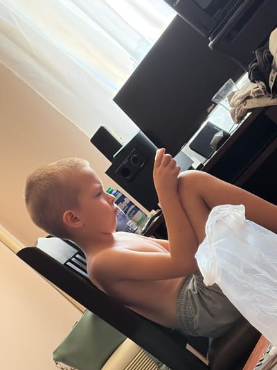 A low-angle shot shows a young child, likely a boy, crouched and looking downwards. He is shirtless and wearing light gray shorts. His arms are wrapped around his knees, and he appears to be holding a crinkled white plastic bag. The child's face is turned to the side, and his expression is neutral.
The background is dimly lit and suggests an indoor setting. To the left of the child, a dark surface, possibly a desk or shelf, is visible. On this surface, there are various objects including a black electronic device with circular buttons, a white plastic bag, and some clothing items draped over it. Further left, a portion of a window with sheer white curtains is visible, letting in soft light, indicating it might be daytime. Behind the child, a dark furniture piece with a striped pattern can be seen.
The scene is cluttered, with the child positioned between a dark electronic device and a patterned surface. The lighting is low, with the primary light source appearing to come from the left side, illuminating the child's skin and the plastic bag. No text is clearly legible in the image, although a logo-like marking is faintly visible on the dark surface to the left. The overall impression is of a casual, possibly relaxed moment indoors. The orientation of the photo is rotated.