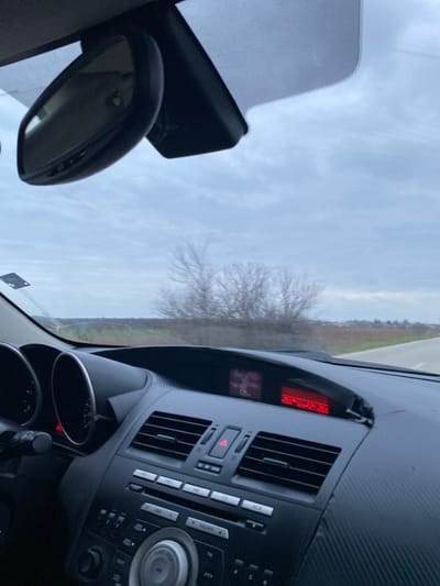 The image depicts the interior of a vehicle, viewed from a passenger's perspective through the windshield, revealing a rural landscape in Sredkovets, Bulgaria, at approximately 23:42:26. The upper portion of the frame shows a grey, overcast sky. Below this, a cluster of bare, deciduous trees is visible in the middle distance. To the right, a paved road recedes into the background, flanked by brown, uncultivated fields. Distant structures indicative of a small settlement are discernible on the horizon.

The immediate foreground reveals the vehicle's dark-toned dashboard and central console. A black-framed rearview mirror is mounted overhead. The dashboard features horizontal air vents, an activated red hazard light button, and two digital displays. The left display shows numerical readings including "7.0" and "126 km" (partially visible), while the right display prominently shows the time "23:42:26" in red numerals. A black panel with a carbon-fiber-like texture surrounds these displays. A portion of the steering wheel and the vehicle's left gauge cluster, featuring an illuminated orange-red needle, are visible on the far left. No occupants are explicitly visible. The vehicle appears to be in motion.