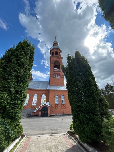 The image shows an exterior view of a brick church with a bell tower in Medvedevo, Russia. The church has a grey roof, arched windows, and a golden dome. A large black double door serves as the entrance. Two tall, dense evergreen trees frame the foreground, with a paved walkway leading towards the church. The sky is partially cloudy with the sun visible and casting rays. The ground in front of the church is asphalt.