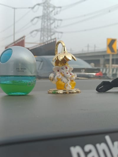 This media file presents a close-up view from the dashboard of a vehicle in Noida, India, looking out onto a road. In the foreground, prominently displayed on the grey dashboard, are two main objects. On the left, a spherical car air freshener, silver at the top and transparent at the bottom, holds a bright green liquid. Visible text on its side reads "05/2024" and "TB022," likely an expiry or manufacturing date and a batch code. To its right, a small, ornate idol of Lord Ganesha sits. The deity is depicted in white and yellow, richly adorned with small, sparkling rhinestones or beads, and is seated beneath a golden, flower-shaped canopy that arches over him. On the far right of the dashboard, a small black fabric object, possibly a strap, is partially visible. A partial white word, "nank," is also faintly visible on the bottom right edge of the dashboard.
The background reveals a hazy, overcast sky, suggesting a cloudy day or possibly significant air pollution. In the distance, a large electricity pylon with multiple power lines stands against the sky. Below it, an elevated road or railway line stretches across the mid-ground. On the right, a yellow road sign with a black chevron arrow pointing to the left indicates a sharp curve ahead. The scene depicts an urban or peri-urban environment with typical infrastructure found in cities like Noida, India. The vehicle appears to be on a highway or major road, either stationary or moving slowly given the foreground clarity.