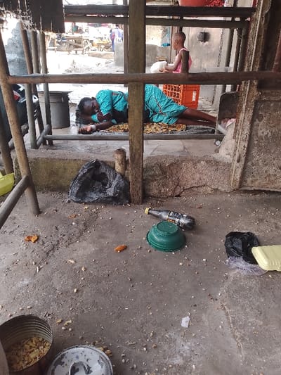 The image depicts a scene within a rudimentary structure, possibly a stall or shelter, featuring both an elevated platform and a concrete ground level.

On the raised platform, a woman wearing a patterned blue-green dress is lying on a mat, appearing to be asleep or resting. Her arm is bent, and her hand is near her head. Further back on the same platform, a young child in a pink top stands, looking away from the camera towards the distant background, possibly holding a white object. An orange plastic crate is visible behind the child. The structure itself consists of sturdy, possibly rusty, metal bars and wooden posts, suggesting a makeshift or functional construction.

The ground level below the platform is made of concrete and is scattered with various debris, including what appears to be food scraps or remnants. Prominent objects on the ground include a black plastic garbage bag, an upside-down green plastic bowl, a dark bottle (likely a beverage, with partially visible but illegible white and yellow text), another black plastic bag, and a yellow foam container. In the bottom-left corner, a metal tin or pot containing light-colored fragments, possibly grains or food, sits with its lid nearby.

In the distant background, beyond the structure, a bustling street or market scene is visible, with faint figures of people and stalls, indicating an active public area. The lighting suggests it is daytime. The overall setting appears informal and lived-in, consistent with a busy, informal urban or semi-urban environment in Mgbuoshimini, Nigeria.