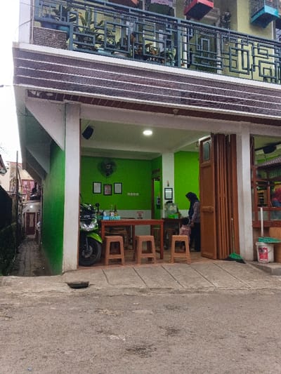 A two-story building with a green-painted storefront open to the street. Inside, a green wall with a ceiling fan and framed pictures is visible. A wooden table and stools are set up, and a green motorcycle is parked inside. A woman in a hijab stands near the counter with a child. The building's upper floor features a balcony with plants and decorative metal railings. The street pavement is visible in the foreground.