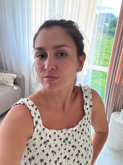 The image is a medium shot selfie of a woman in her late 20s or early 30s with her hair pulled up. She is wearing a white smocked dress with a black palm tree pattern. The woman has light skin with freckles and dark hair. She is looking directly at the camera with a neutral expression. Behind her, to the left, is a gray sofa with a decorative pillow. To the right, a window with sheer white curtains on the left and pinkish-brown drapes on the right reveals a green, grassy outdoor area with some structures visible in the distance, possibly in the city of Nizhnyayarkeyevo, Russia. The lighting suggests it is daytime, with sunlight filtering through the window.